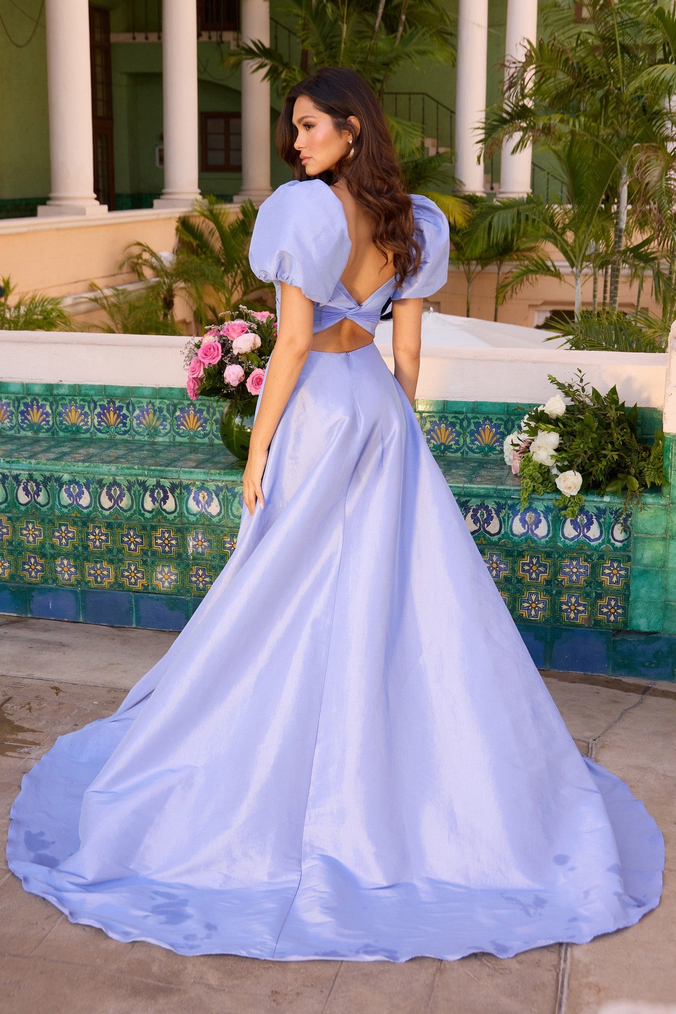 Woman stands in a courtyard, surrounded by greenery and colorful tiles, wearing the Puff-Sleeve Long Prom Dress: Ava Presley 42269—a light blue A-line gown with puff sleeves and an open back.